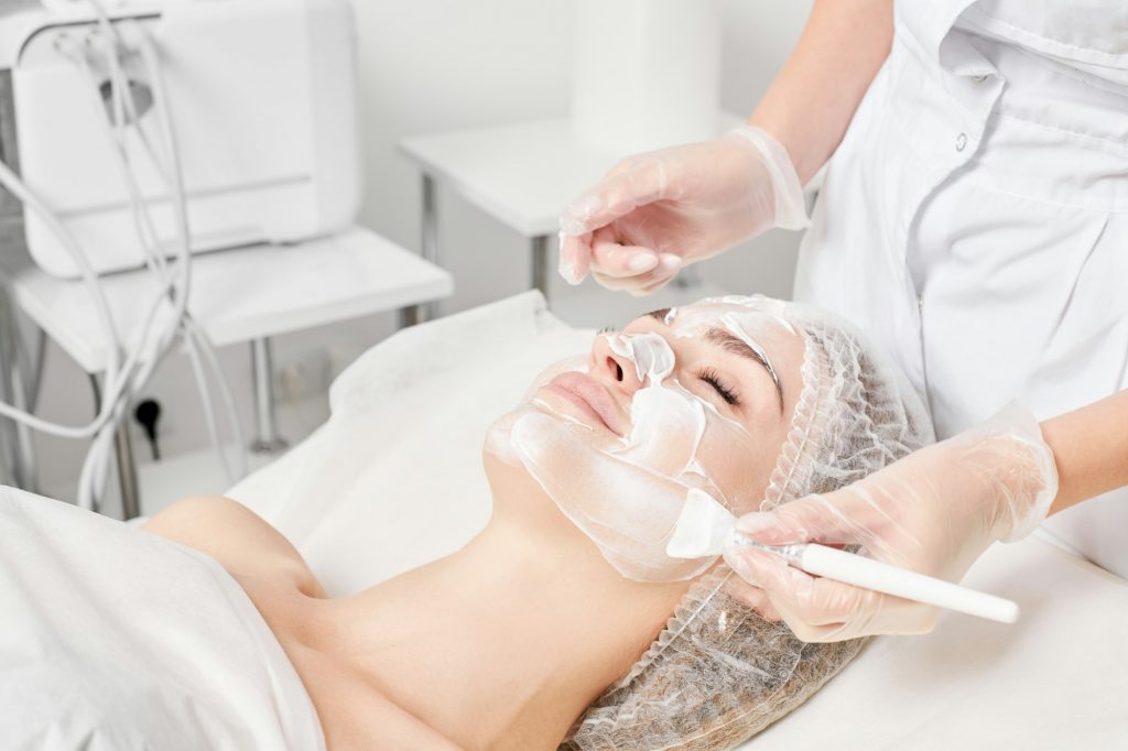 Beautician applies cream mask on woman face for rejuvenation face skin, procedure in beauty salon