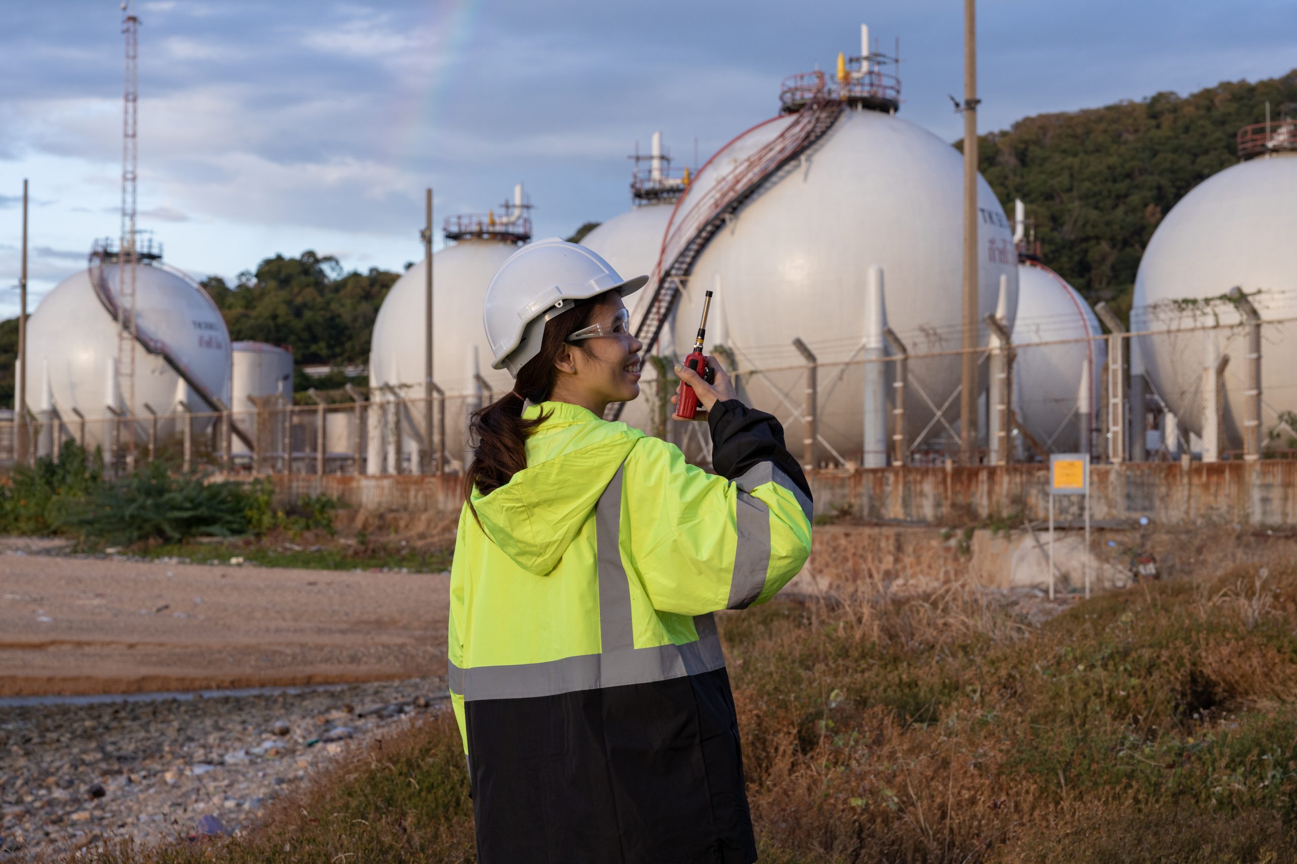 Female engineer in safety gear using a walkie-talkie near large spherical gas storage tanks at an industrial site.
